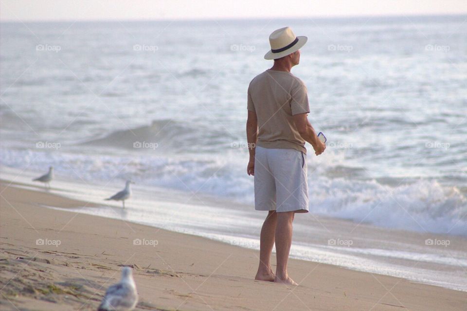 A man in a hat walking along the beach at dawn. 