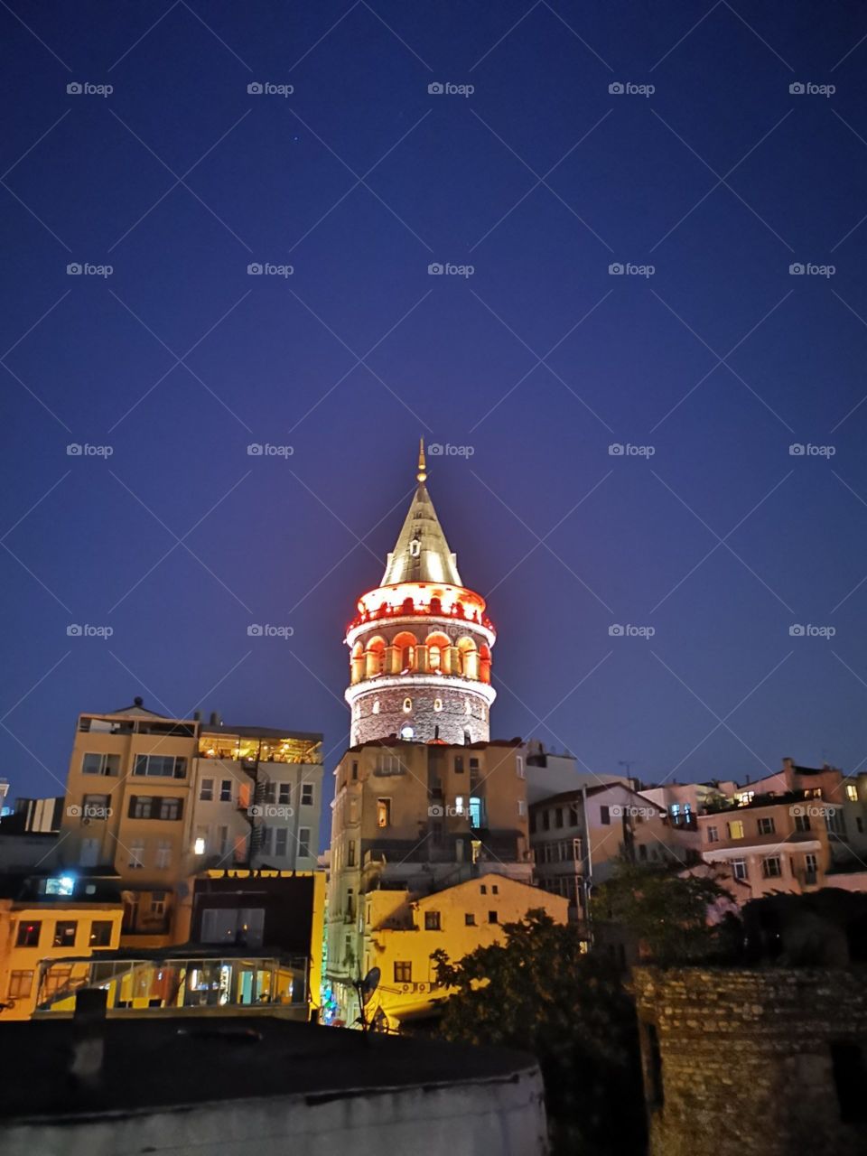 The old tower artificially iluminated with red and white lights. And iluminated human houses below the tower. With beautiful blue cloudless sky in background.