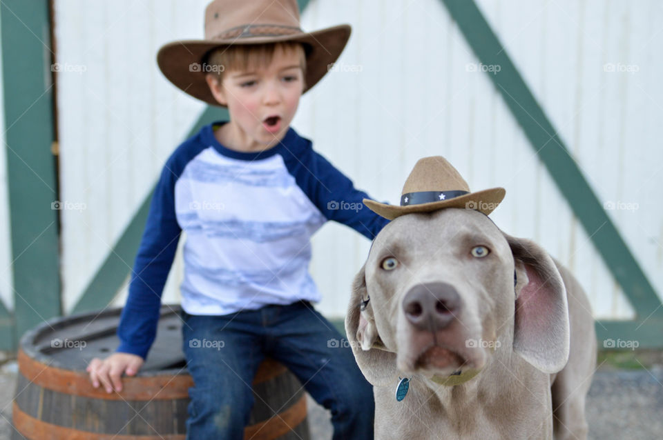 Young boy chasing after his Weimaraner dog, both wearing cowboy hats in front of a barn door outdoors