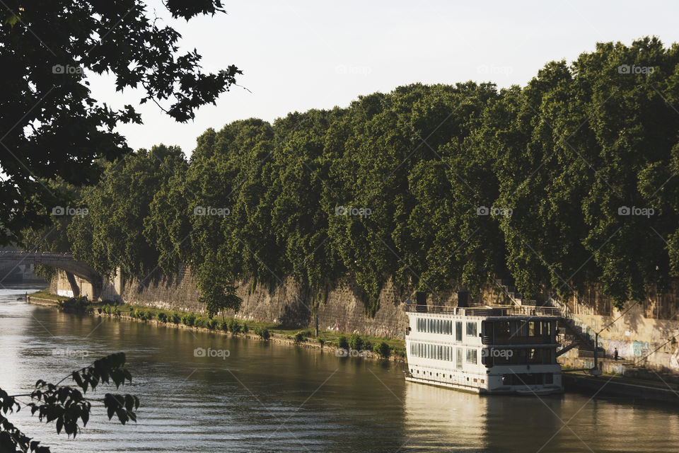 A landscape portrait of a big river in Rome, Italy. on the river their is a big olg ferry boat. the river is surrounded by trees.