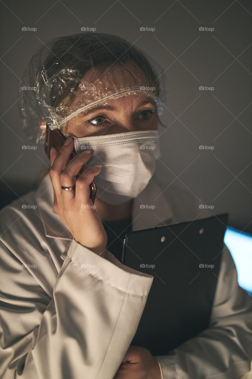 Doctor making a phone call. Hospital staff working at night duty. Woman wearing uniform, cap and face mask to prevent virus infection