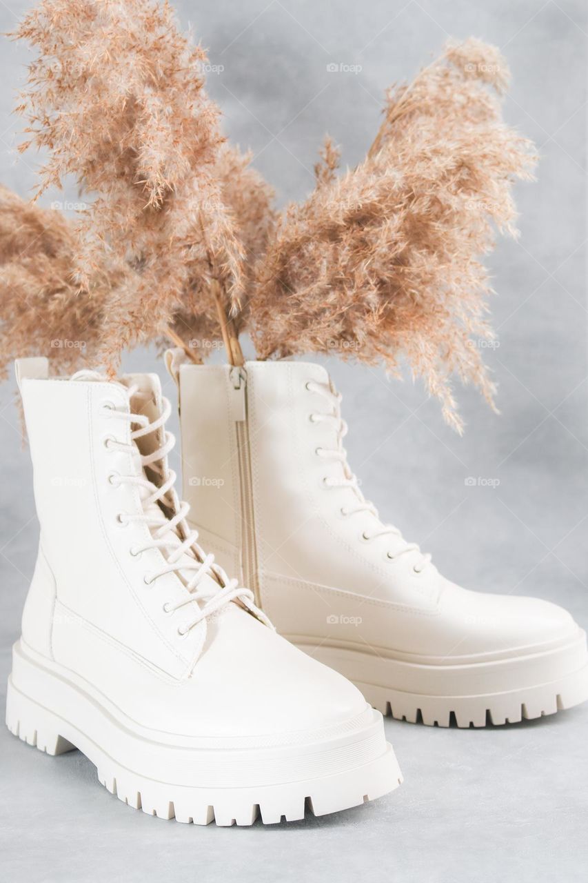 Beautiful white boots with a rough tractor sole laced eco leather with a bouquet of dried flowers standing on a gray cement background,side view. The concept of fashionable women's shoes.