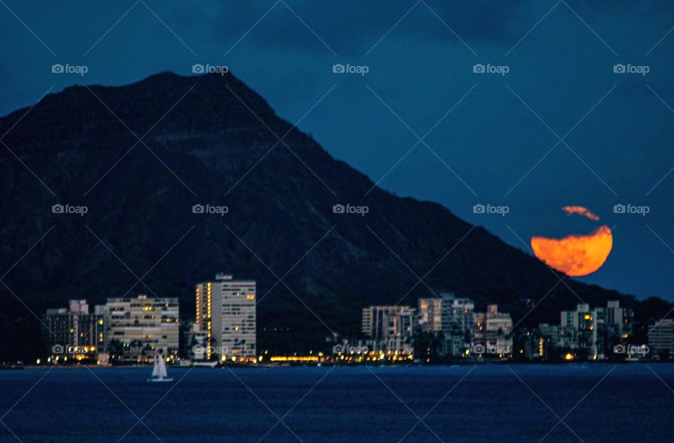 A bright orange moon rises behind Diamond Head, a prominent peak in Honolulu