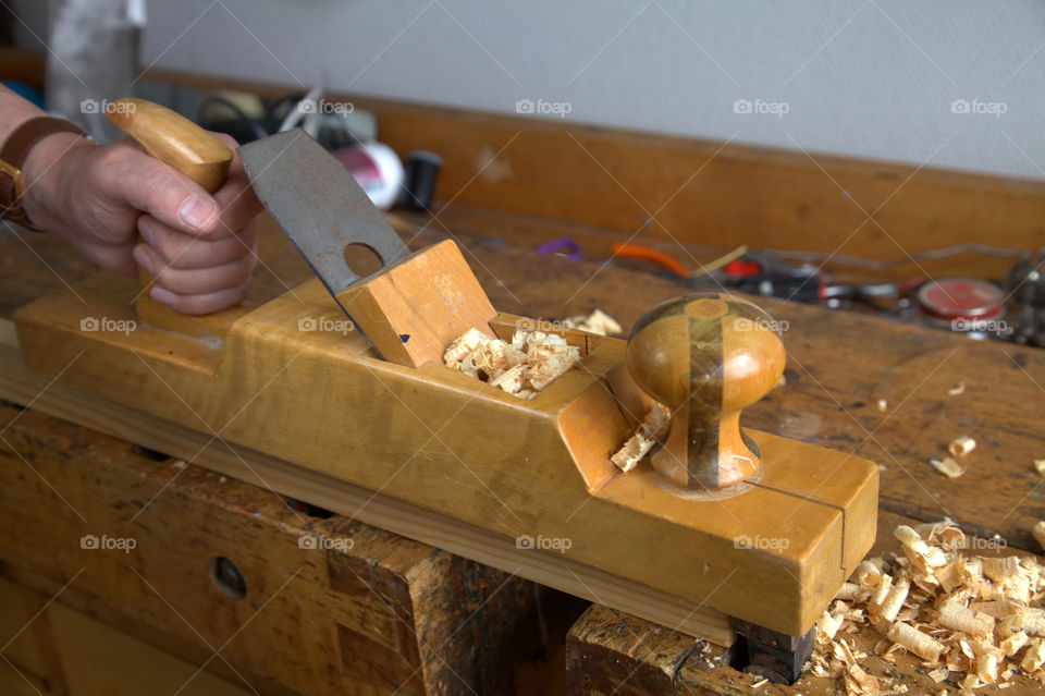 Hand of an old carpenter holding old wooden planer on a workshop table with wood chips, other instruments, jars and cans around.