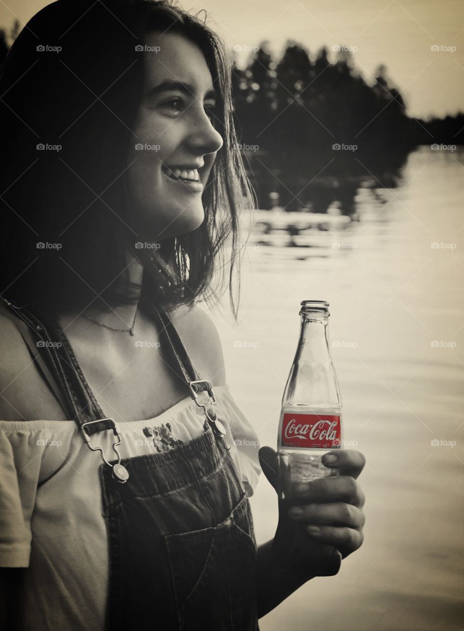 Girl drinks a coke while socializing on a lake. Black and white, Coca Cola label visible color