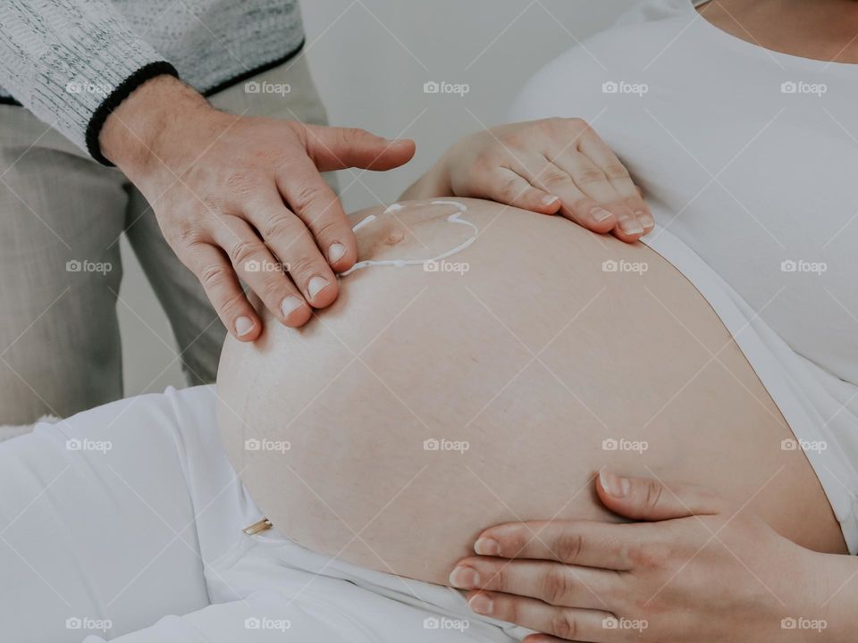 A close-up side view of a man's father's hand massaging and smearing cream on the belly of his pregnant wife lying on the bed during the day.