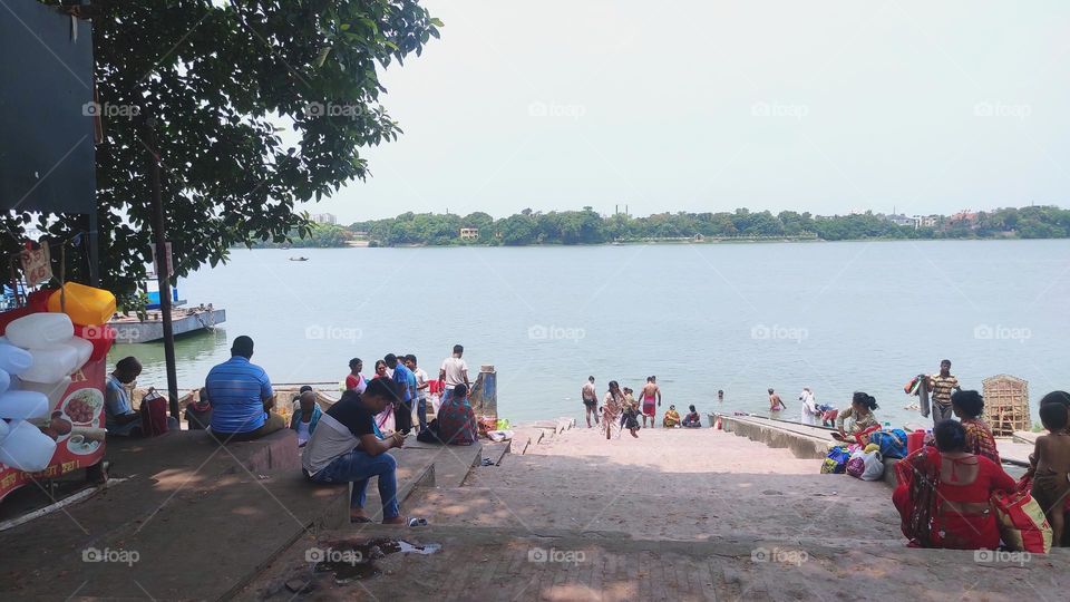 This is a photo of river side of Ganga or Ganges Panihati of West Bengal. Some people are resting and some people are taking bath. Spreads tree shadow is showing in the photo.