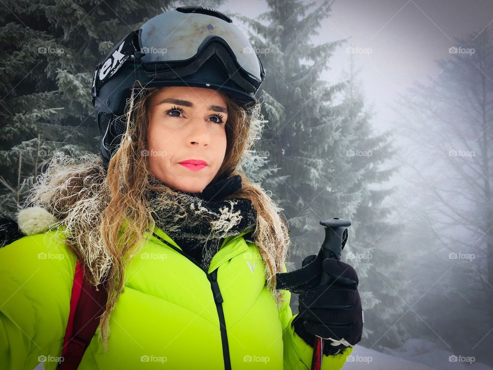 Portrait of woman wearing Uvex ski glasses and Uvex ski helmet and holding ski poles with pine cone trees covered in snow in the background