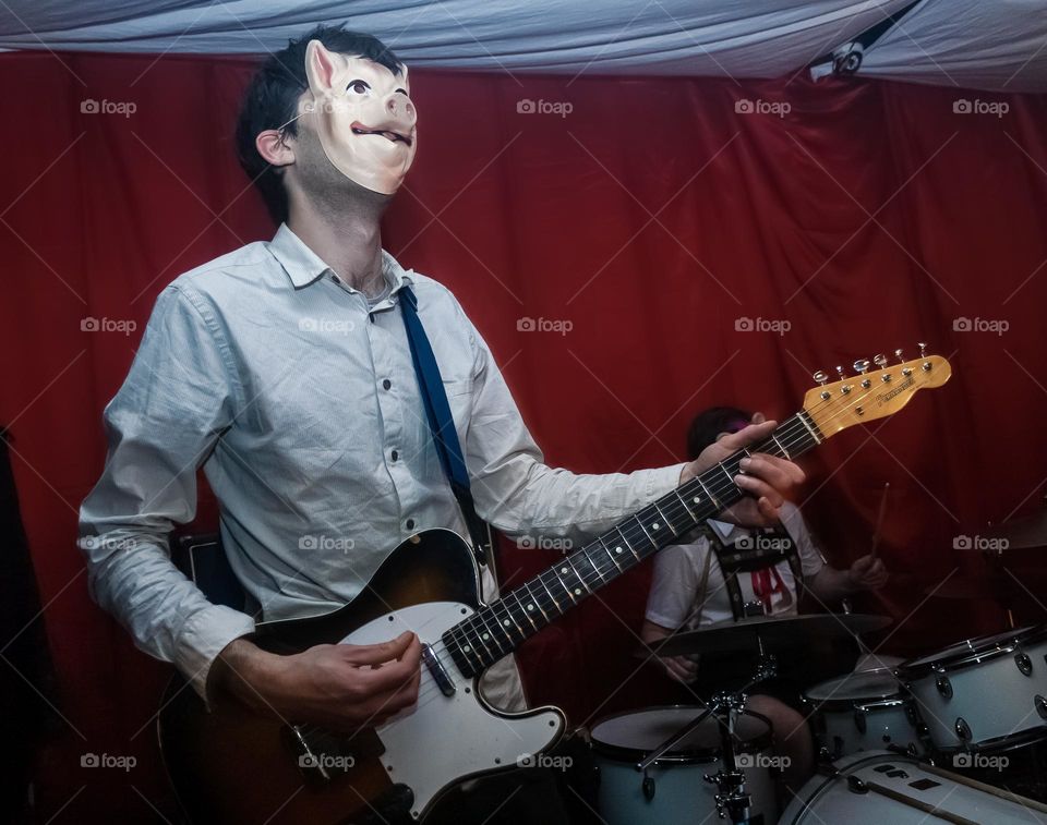 A man wearing a pig mask plays guitar with a drummer behind them