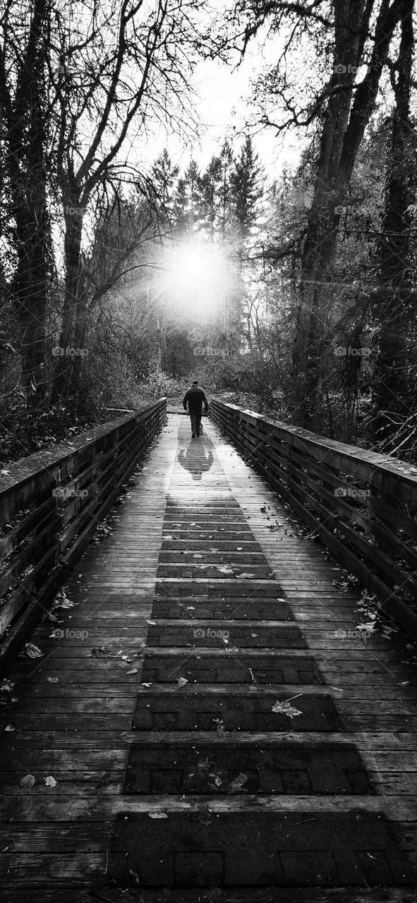 filtered black and white photo of a man walking on a trail bridge in Oregon toward the bright light casting a shadow behind him