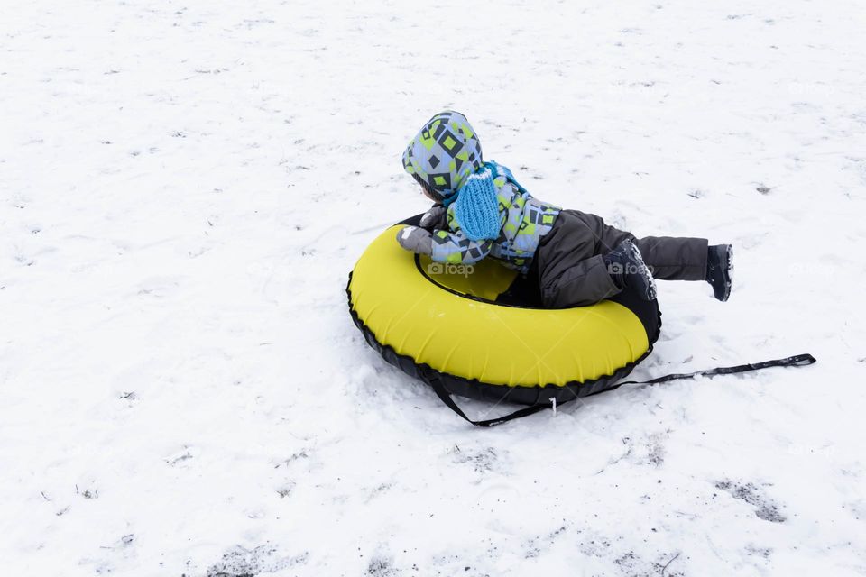 A small, carefree boy walks in the white snow in winter and rides a tubing in the park, near trees in the snow.