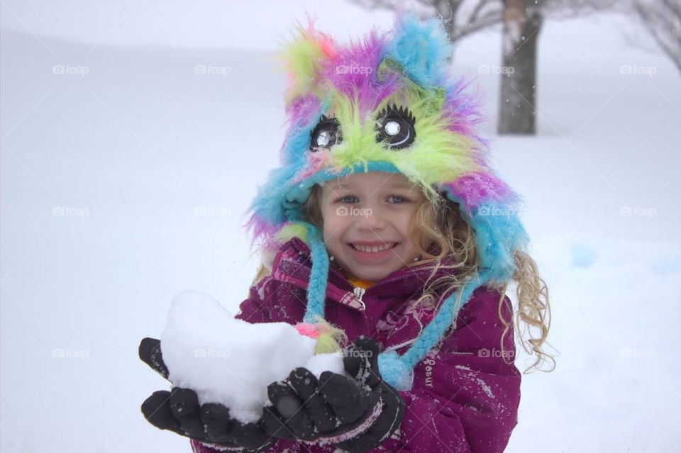 A sweet little girl in a colorful hat, holding a giant chunk of snow that she plans to throw at her brother.