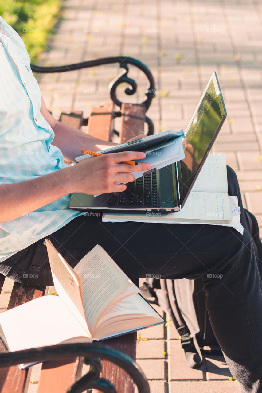 Student working on a laptop using books and notes sitting on a bench in a park. Young boy wearing a blue shirt and dark jeans