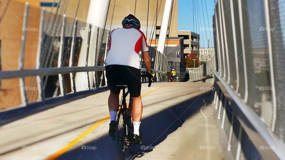 A man on a bicycle and a woman in the distance jogging in an urban setting on a pedestrian bridge staying in good shape