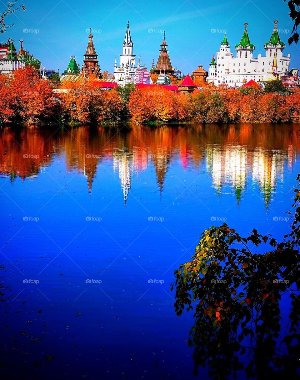 Autumn landscape.  On the bank of the river, among the autumn trees, the domes of the towers are visible.  Trees and domes of towers are reflected in the dark blue water.  In the foreground is a birch branch with green and yellow leaves.