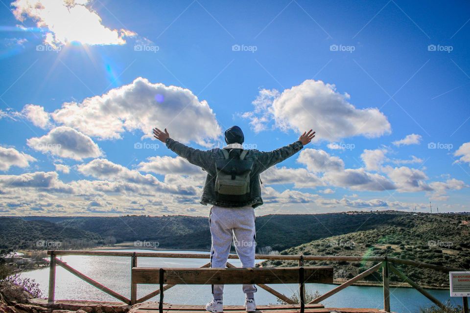 a man with open arms on top of a hill of a lake with sunny blue sky and clouds