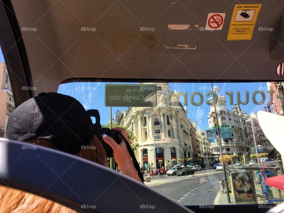 Person seen from behind sitting in the front on a tour bus taking pictures out on the street 