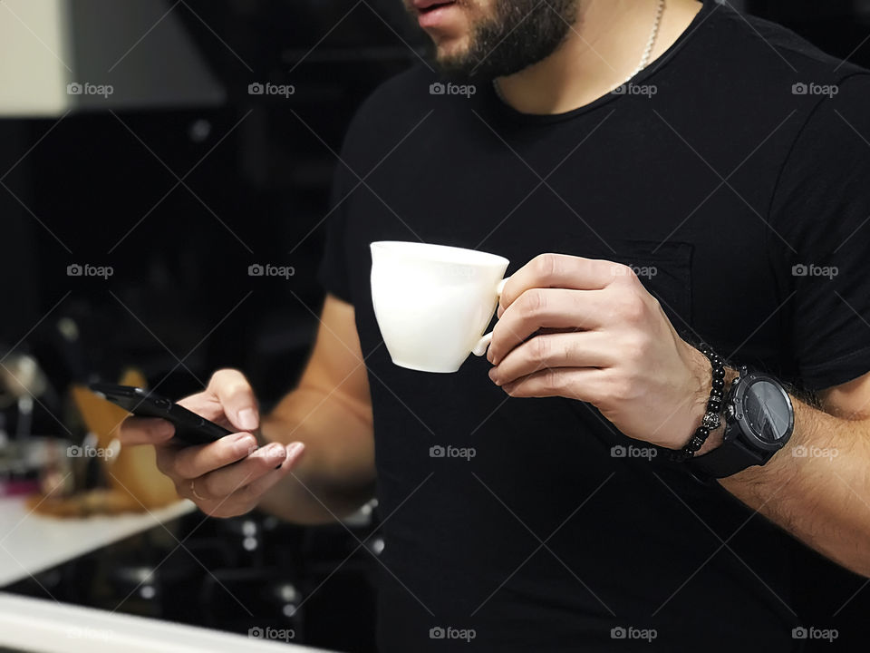 Young modern man wearing smart watches and using mobile phone while drinking morning coffee in the kitchen at home before going out 