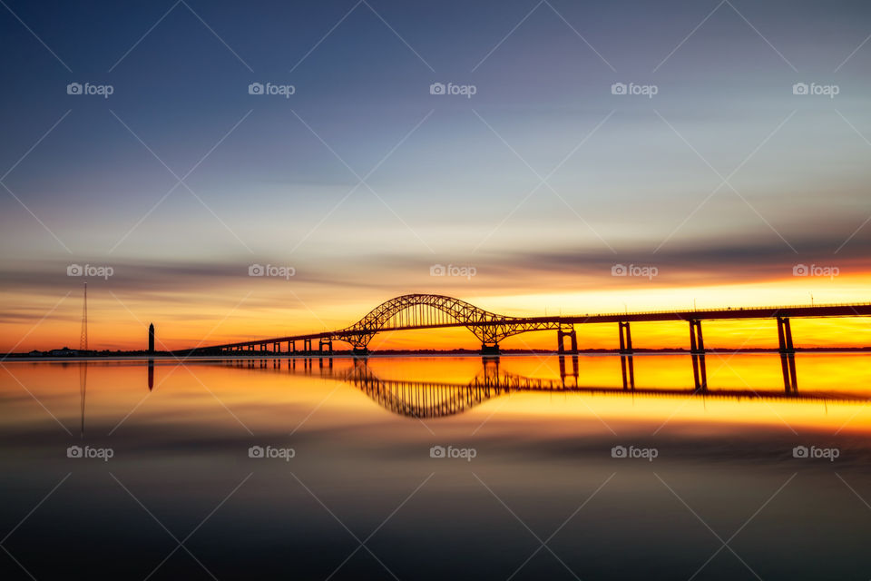 Silhouette of a long arched bridge stretching across a calm body of water at sunset. Clouds and bridge reflected perfectly in the water