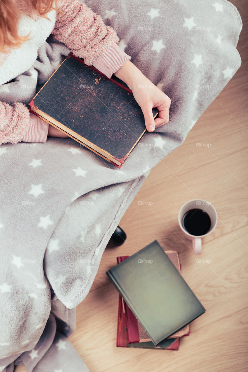 Girl enjoying the reading a book and drinking coffee at home. Young woman sitting on a chair, wrapped in blanket, holding book, relaxing at home. Portrait orientation. View from above