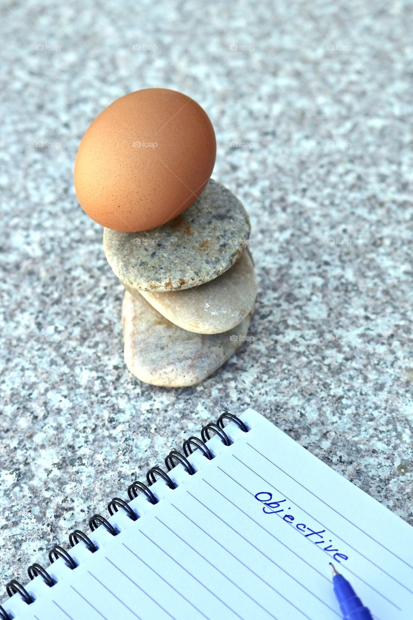 Brown egg balancing atop smooth stone rock stack on marble surface, with spiral notebook, objectives handwritten on open page, concept life, business, ideas, creativity endurance or stability and planning and organisation