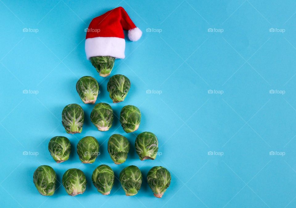 Brussels sprouts lined up in the shape of a triangular Christmas tree with a mini Santa Claus hat on the left on a blue background with a copy of the space on the right, flat lay close-up. Holiday Vegetables Concept.