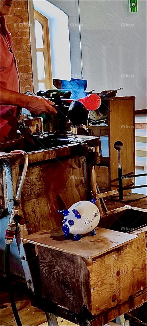 A piece of red hot glass s giving off smoke as its being manipulated at the end of a long thin pipe in this workshop in “Freyung”, Bavaria, Germany. 2023. Hypnotic Productions