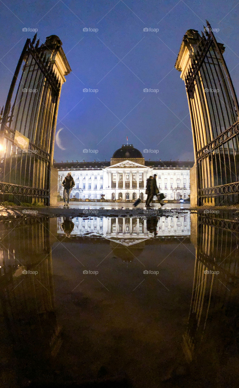 Royal Palace of the Belgian king as seen through the gates of the king’s parc and reflected in the water of a puddle during the rain at night, in Brussels, Belgium