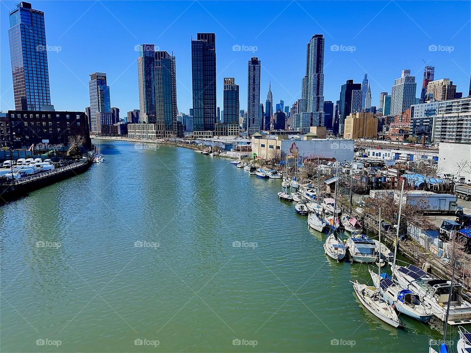 This is a magnificent view of “Newtown Creek” with its various boats from the “Pulaski Bridge” that connects LIC, Queens to “Greenpoint”, Brooklyn. In the distance we see “Manhattan” incl. the “Empire State Bldg”. 2024. Hypnotic Productions