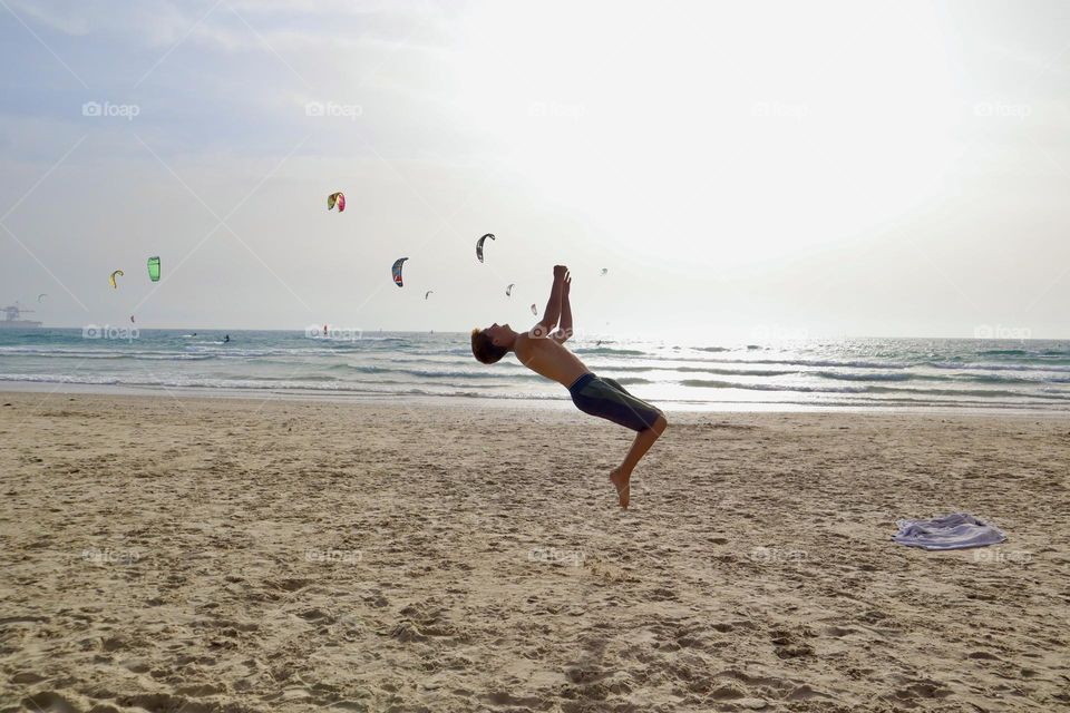 Kid doing somersault on the beach with the sea and kite surfers on the background 