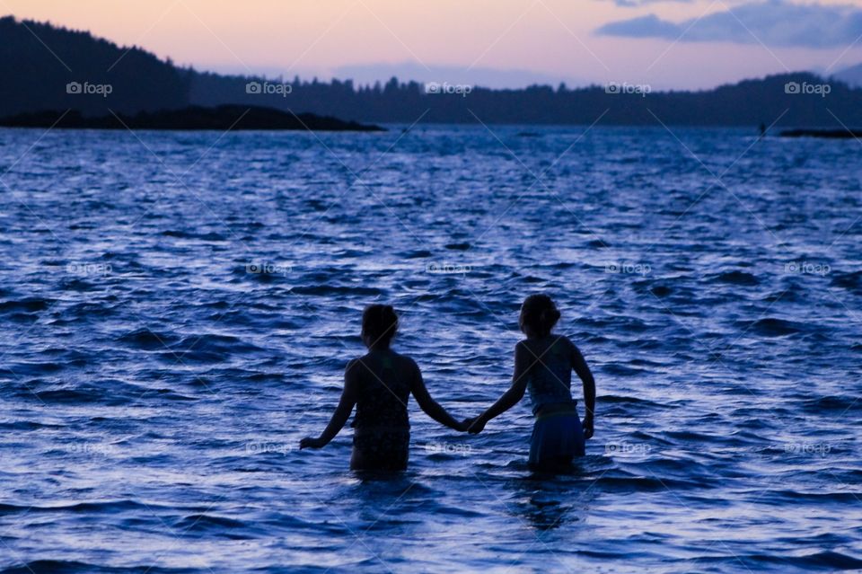 My 2 daughters, bravely inch hand-in-hand into the cold Pacific water to play in the last remnants of sunset.  The black silhouettes of their bodies & the coastal tree line are a stark contrast against the inky blue water and the pink & purple sky.👥