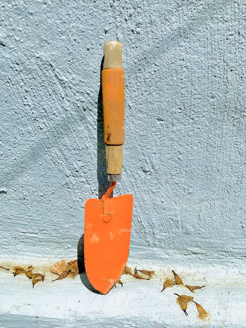 A small orange hand shovel for gardening against a light blue wall. In a background garden.