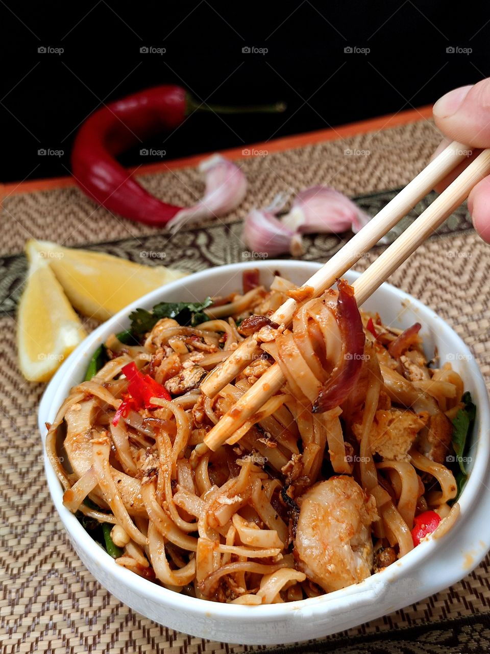 Thai noodles.  A white plate contains noodles, squid rings, shrimp and spices.  A female hand holds noodles with wooden chopsticks.  Next to the plate are garlic wedges, lemon wedges and red chili peppers