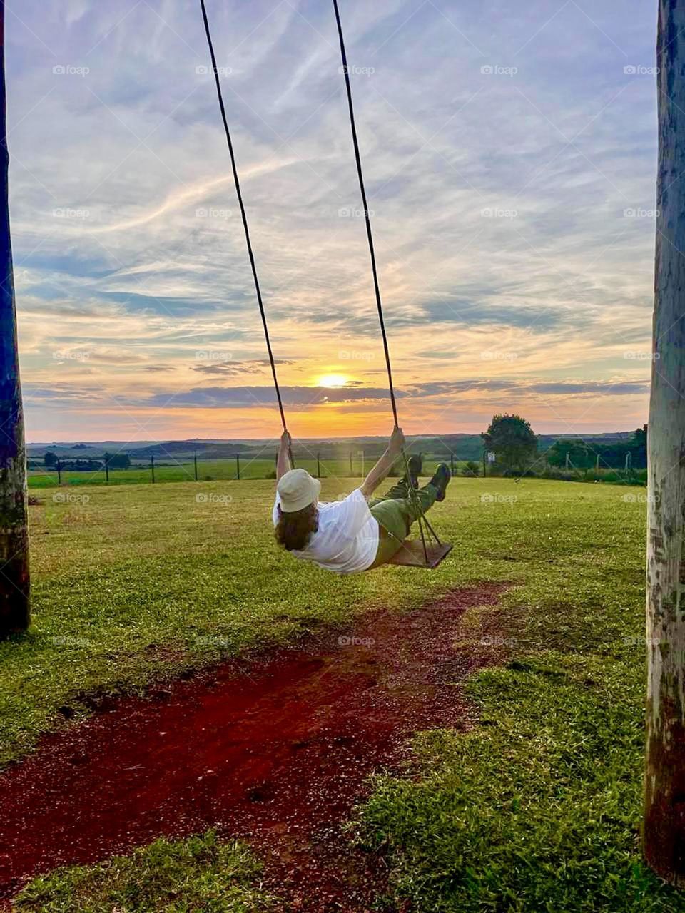 A person playing on a swing under a dazzling sky during sunset, with colorful clouds and a green horizon in the background, capturing a moment of happiness and freedom.