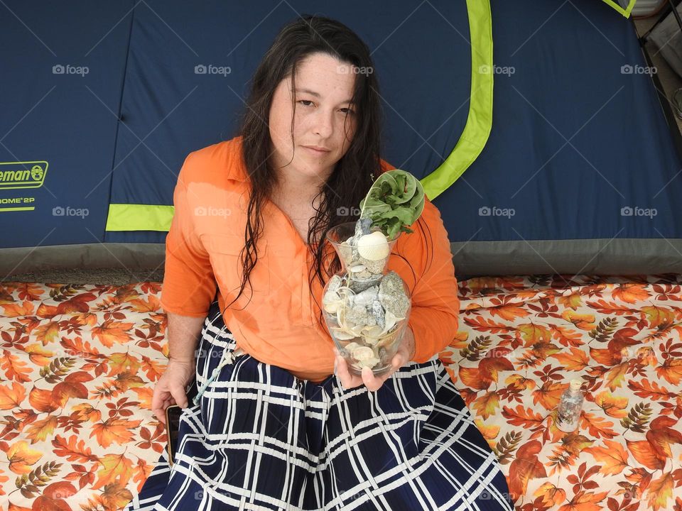 Autumn fall decor season during camping, woman sitting on a step on a cloth with orange leaves with blue and orange clothes in front of a blue and green tent and holding a seashells vase in memory of the past season from summer time.