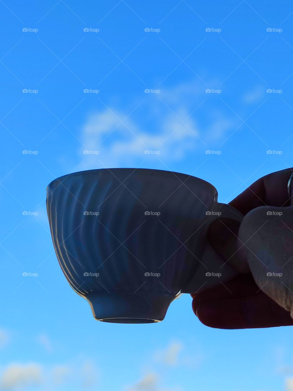 A woman's hand holds a white cup of coffee on a background of blue sky with white clouds.  White clouds like steam over a cup