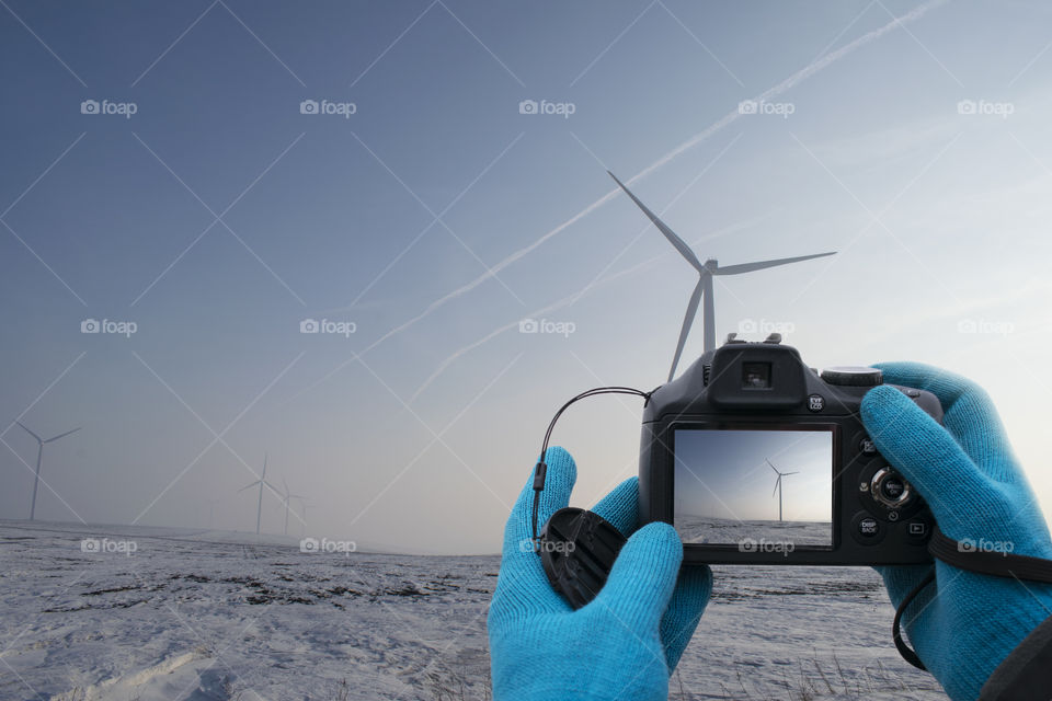 Compact camera in hands with blue gloves take picture of wind turbines in winter season