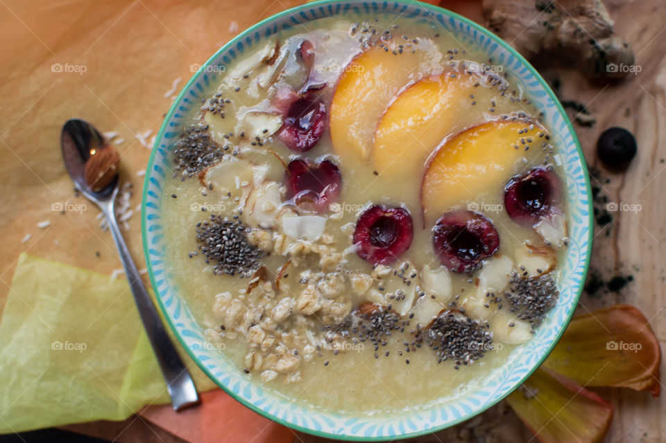 High angle view of fresh fruit smoothie bowl with pretty sliced peach, cherry, almond, chia seed, coconut and granola on table with spoon 