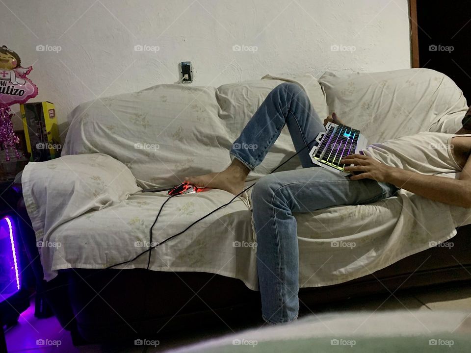 Man lying down on a sofa, while playing a video game with a keyboard and mouse with his feet in a funny way.