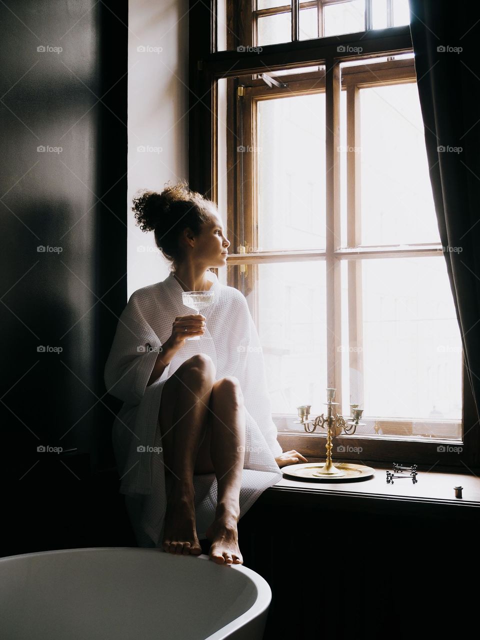 Young beautiful woman with curly hair wearing white bathrobe sits on the window in the bathroom and drinks champagne, portrait of woman, looking at window, lifestyle, at home, relax.