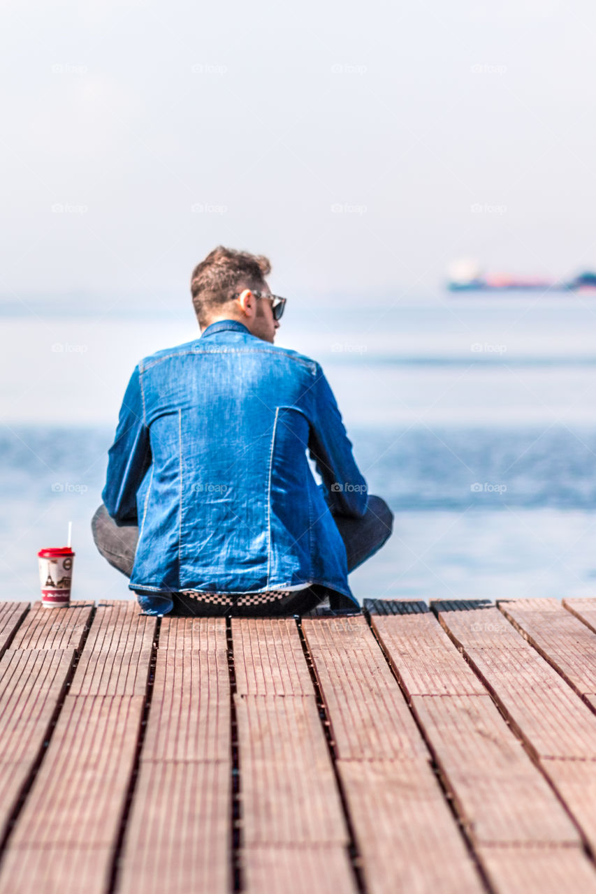 Man Wearing Denim Jeans Clothes Sitting On A Dock And Looking At Blue Sea
