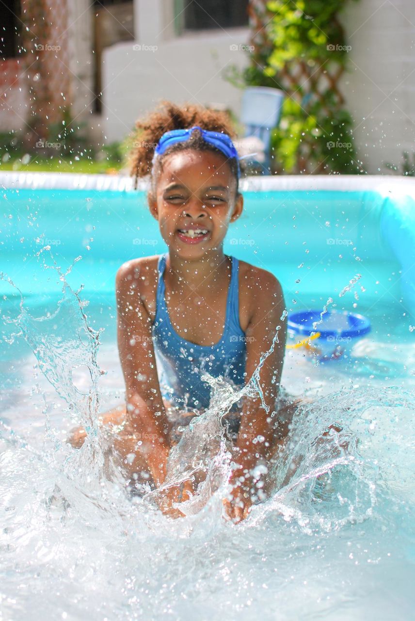 Girl of mixed race enjoying the refreshment of water in a swimming pool on a hot summer day, together with her little sister (family, fun, summer, water, blue, swimming suit, splash, hot, enjoy, play, outdoors)