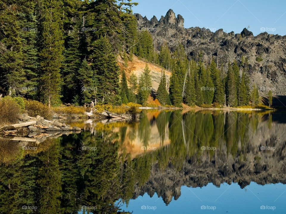 Jagged and rugged hills of hardened lava rock as well as evergreen trees and a rich blue sky reflect in the smooth waters of Devils Lake in the mountains of Central Oregon on a sunny fall day.