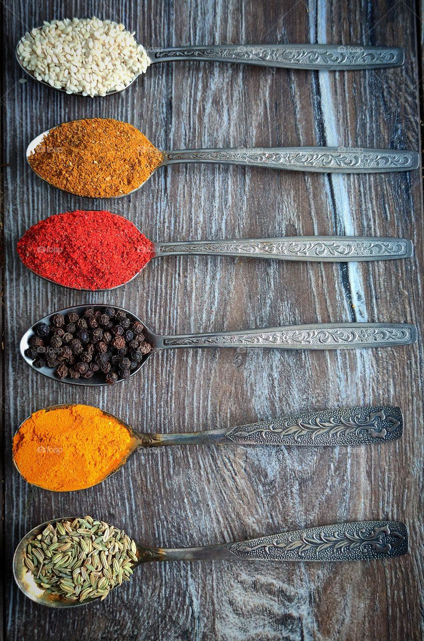 On a wooden surface are six metal spoons with spices: green fennel, yellow turmeric, black peppercorns, red paprika, orange curry and white sesame. Spice color contrast. Wood and metal contrast