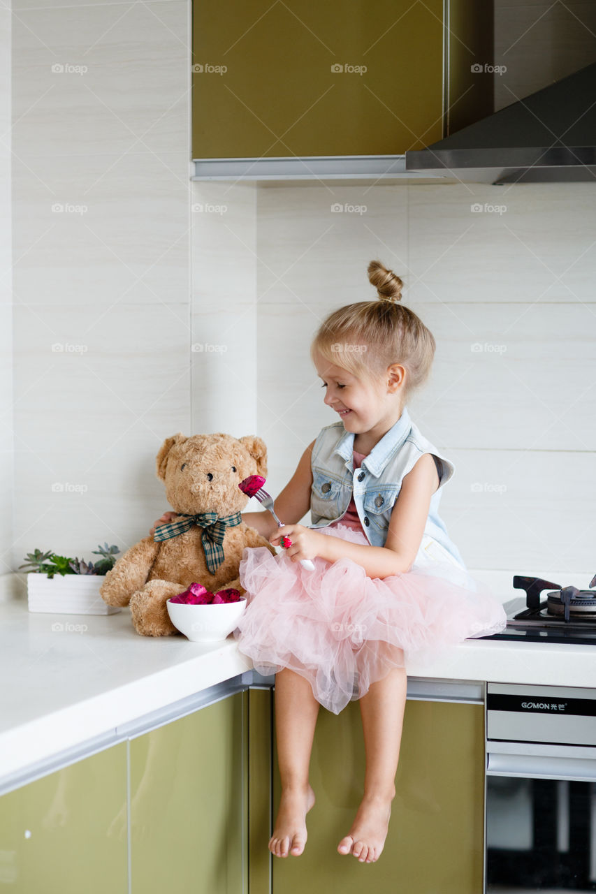 Happy little girl with blonde hair and teddy bear on the kitchen 