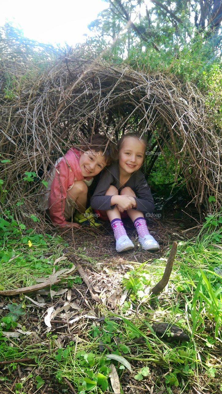 two kids a boy and a girl sitting together under a bush shaped as a little hut or cubby house