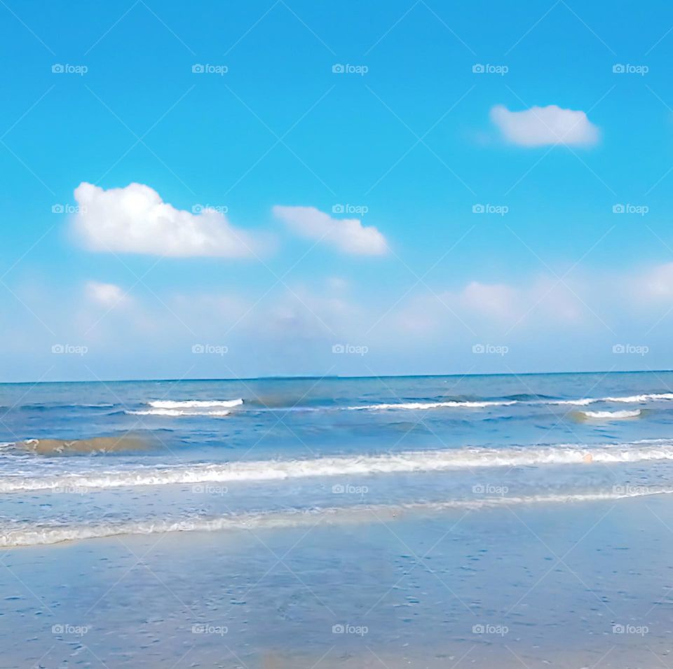 The image showcases a tranquil beach scene with gentle waves rolling onto the shore. The sky is a brilliant blue with a few fluffy white clouds scattered across it, adding depth to the scenery. The horizon line is clearly visible, where the sea meets
