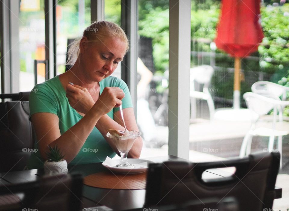 Portrait of a beautiful young and sad blonde woman sitting alone at a table in a cafe and eating ice cream with her arms crossed, close-up side view. The concept of sad people, lonely women.