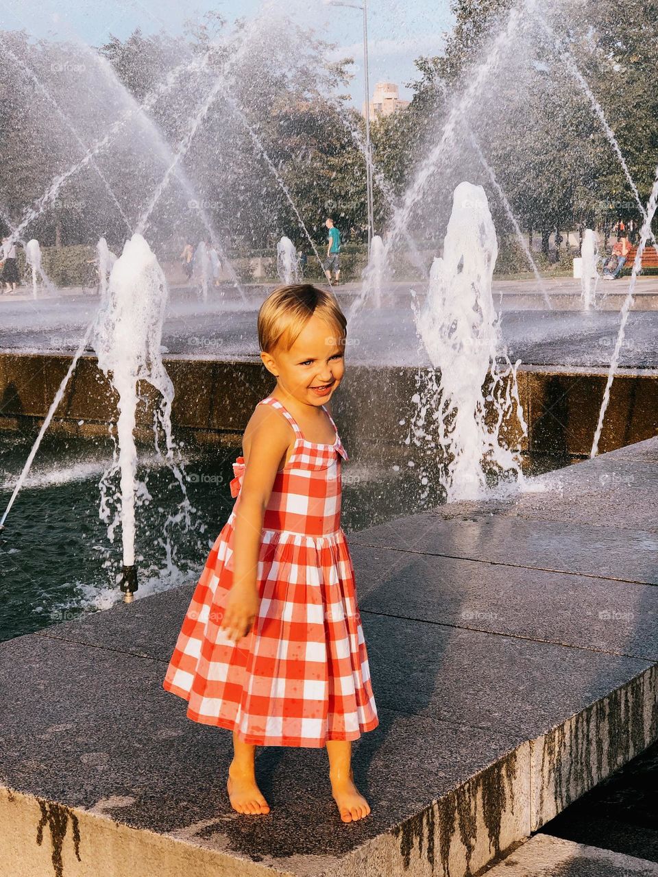 A cute little smiling blonde girl walking near the fountains on a summer sunny day, portrait of child 