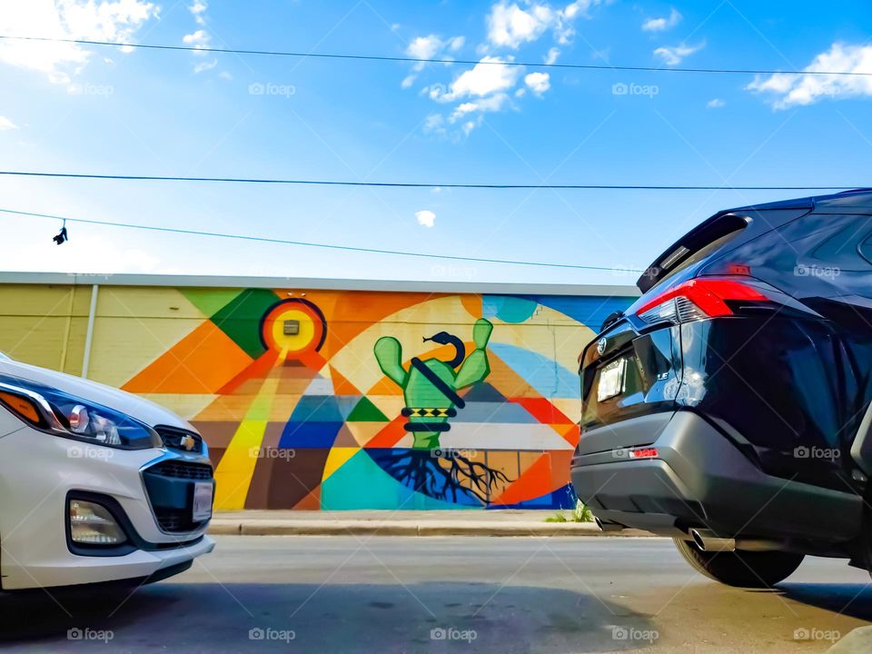 From the ground, view of colorful street art on a building wall across the street, between two parked cars.